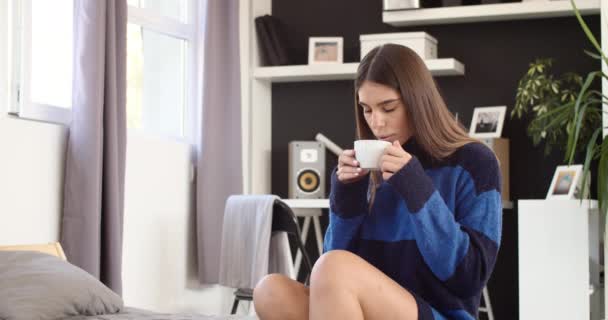 朝寝室に座ってコーヒーを飲む青いセーターの魅力的なブルネット — ストック動画