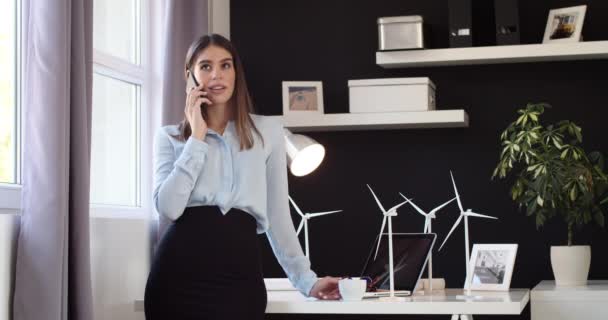 スマートフォンや飲酒コーヒーを使用して オフィスに立って正式な摩耗に身を包んだ魅力的なビジネス女性 テーブルの上に風車モデルとラップトップです 持続可能な開発の概念 — ストック動画