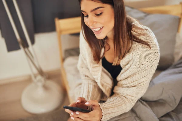 Visão superior de atraente morena caucasiana vestida com suéter bege sentada na cama no quarto e usando telefone inteligente . — Fotografia de Stock