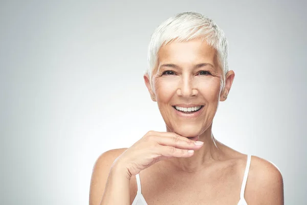 Mooie lachende Senior vrouw met korte grijze haren poseren in de voorkant van de grijze achtergrond. Beauty fotografie. — Stockfoto