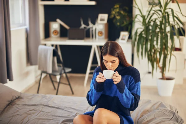 Affascinante bruna in maglione blu seduta in camera da letto al mattino e bere caffè . — Foto Stock