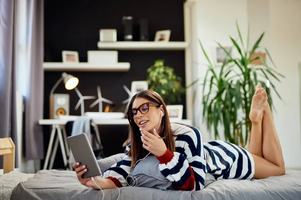 Приголомшлива брюнетка в светрі лежить на животі в ліжку в спальні і використовує планшет. Ранок час . — стокове фото