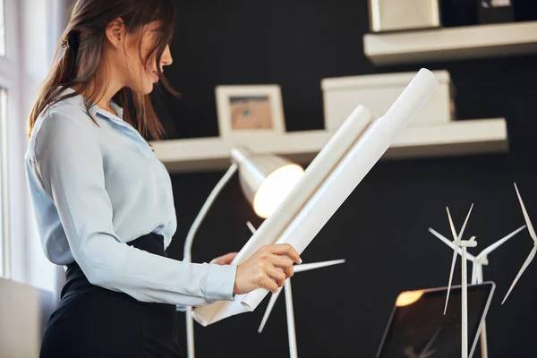 Atractiva mujer de negocios caucásica en camisa y falda de pie en la oficina junto a la ventana y mirando el plano. En el escritorio hay modelos de molino de viento y portátil. Concepto de desarrollo sostenible . — Foto de Stock