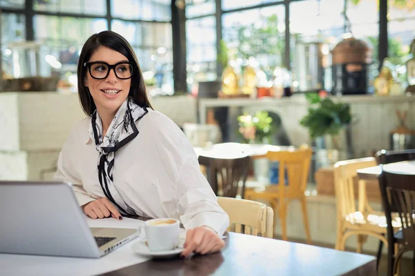 Aantrekkelijke Kaukasische elegante brunette met bril zittend in café, koffie drinkend en wegkijkend. Op tafel ligt een laptop. — Stockfoto