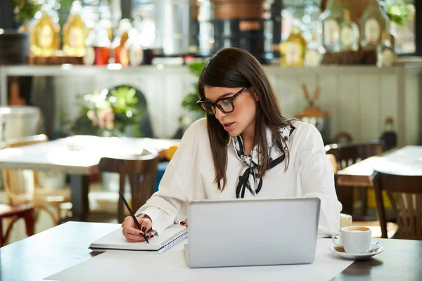 Encantadora mujer de negocios caucásica con gafas sentadas en la cafetería y tareas de escritura en el cuaderno. Delante de ella en la mesa está el ordenador portátil . —  Fotos de Stock