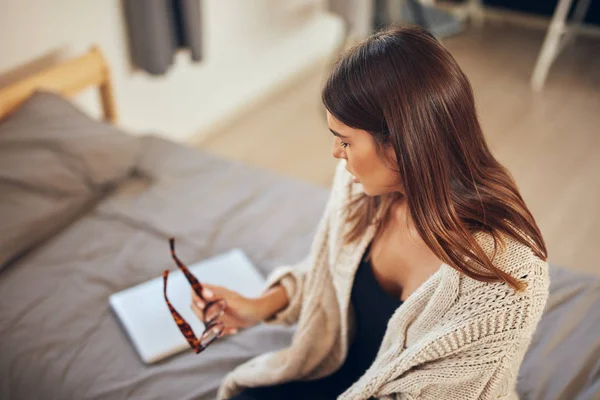 Beautiful slim caucasian brunette dressed in beige sweater sitting on bed in bedroom. — Stock Photo, Image