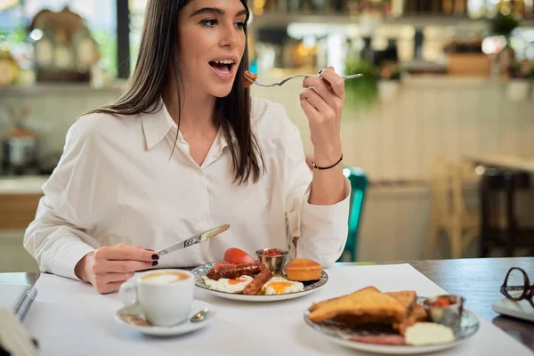 Prachtige Kaukasische elegante brunette zittend in restaurant en worstje etend als ontbijt. Op tafel ligt een laptop. — Stockfoto