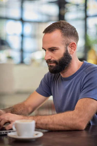 Bellissimo freelance caucasico barbuto seduto nel caffè e utilizzando il computer portatile. Sul tavolo accanto al computer portatile è una tazza di caffè . — Foto Stock