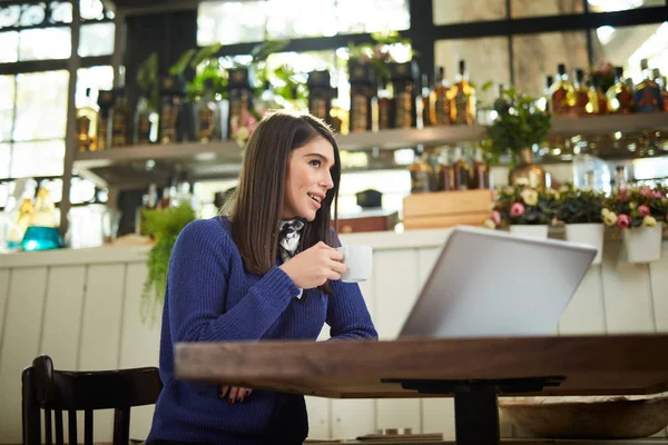 Atractiva morena caucásica en suéter sentada en la cafetería y café molido. En la mesa está el portátil . — Foto de Stock