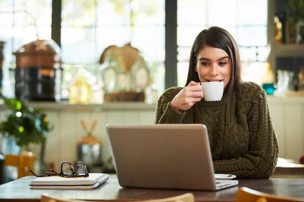 Morena caucasiana atraente em suéter sentado no café e bebendo café. Na frente dela está o laptop . — Fotografia de Stock