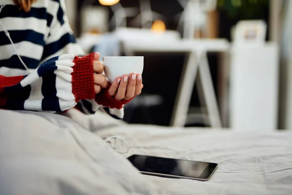 Mujer caucásica en suéter a rayas sentada en la cama, escuchando música sobre la tableta y sosteniendo su café fresco por la mañana . — Foto de Stock