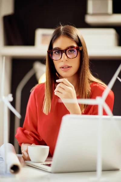 Mooie Kaukasische zakenvrouw gekleed in rode blouse zitten in modern kantoor en driking koffie. Op tafel liggen blauwdrukken, laptop- en windmolenmodellen. Duurzame ontwikkeling. — Stockfoto