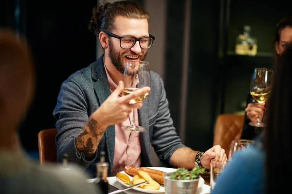Guapo caucásico barbudo hipster hombre con gafas sonriendo y sosteniendo un vaso de vino mientras se sienta con sus amigos en el restaurante . —  Fotos de Stock