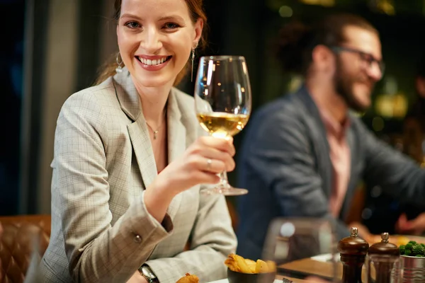 Retrato de hermosa morena caucásica vestida elegante posando con copa de vino mientras estaba sentada en el restaurante con sus amigos . — Foto de Stock