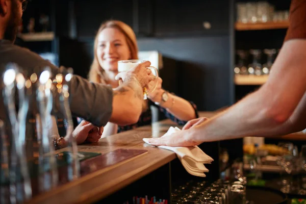 Barman limpando bar balcão enquanto casal inclinado no balcão e fazendo um brinde com cerveja. Foco seletivo na mão . — Fotografia de Stock