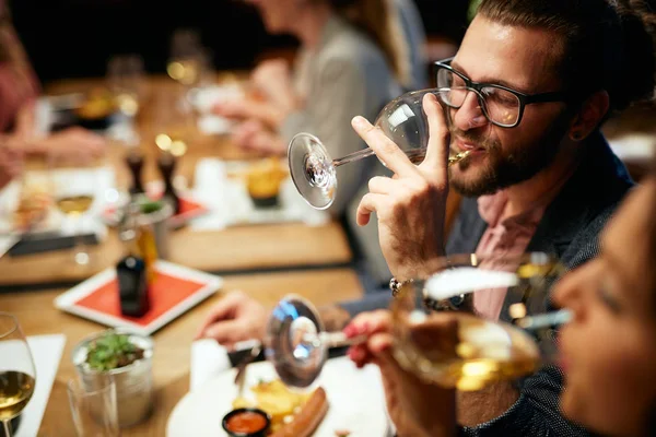 Красивый белый бородатый хипстер в очках, пьющий вино, сидя со своими друзьями в ресторане . — стоковое фото