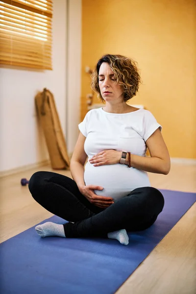 美しい中年妊娠中の女性座っていますマットに自宅で 触手腹と呼吸ヨガの演習を行います — ストック写真