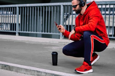 Genç yakışıklı sporcu şehirde uzun bir koşudan sonra çömelmiş, dinleniyor ve akıllı telefonu kullanıyor..