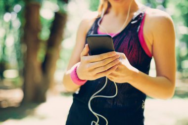Doğada duran ve müzik aramak ya da mesaj okumak için akıllı telefon kullanan genç, fit, beyaz kadın sporculara yakın çekim.