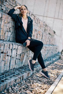 Genç, gülümseyen, zayıf bir bayan koşucu açık havada oturuyor, müzik dinliyor ve koşuya ara veriyor..