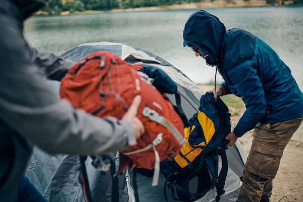Two men on camping trip putting backpacks into tent by the lake on rainy and cold weather.