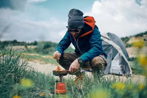 Young handsome man crouching by the tent in nature, looking at beautiful lake and warming up his lunch.