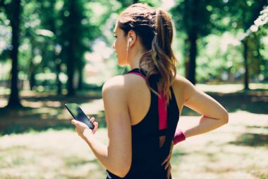 Çekici beyaz kadın koşucunun arka görünüşü doğada duruyor, akıllı bir telefon tutuyor ve ara verirken müzik dinliyor..