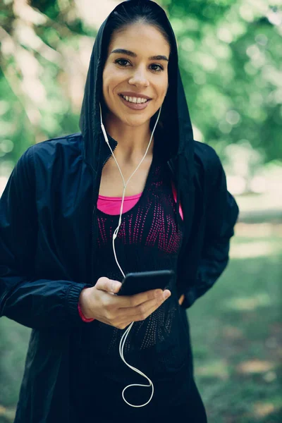 Young attractive caucasian female runner in hooded shirt standing in nature, listening music over cellphone and looking at camera while taking a break.