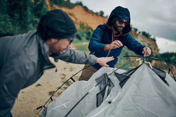Two attractive men disassembling tent by the lake on the camping trip.