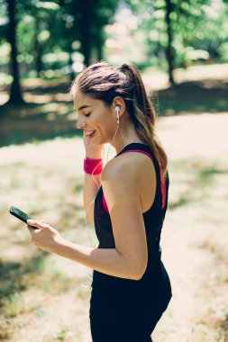 Genç, çekici, fit, beyaz tenli sporcu kadın doğada duruyor ve akıllı telefonu kullanarak koşmak için müzik arıyor..