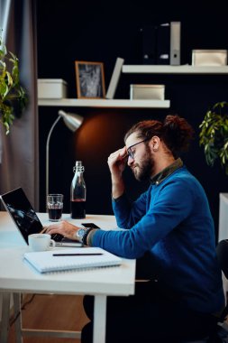 Genç beyaz iş adamı ofisinde oturuyor, dizüstü bilgisayar kullanıyor ve başı ağrıyor..