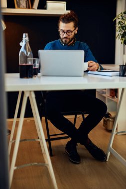 Genç ve çekici sakallı hippi ofisinde oturmuş, kahve içiyor ve akıllı telefon kullanıyor..