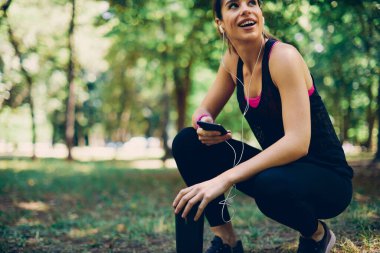 Genç, güzel, gülümseyen sporcu kadın ormanda çömelmiş, ferahlatıcı bir şeyler tutuyor ve akıllı telefondan müzik dinliyor..