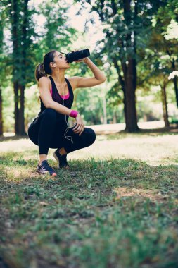 Genç beyaz kadın koşucu ormanda çömelmiş, dinleniyor ve içecek bir şeyler içiyor..
