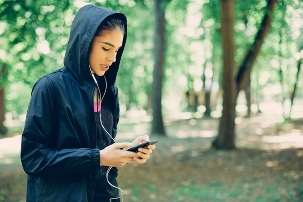 Young attractive caucasian female runner in sportswear standing in forest and using smart phone.