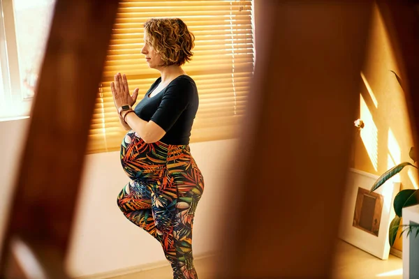 Middle aged fit pregnant woman in sportswear standing in Tree yoga position at home.