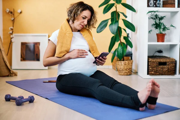 Tired sweaty middle aged pregnant woman sitting at home on yoga mat and using smart phone during corona virus outbreak.