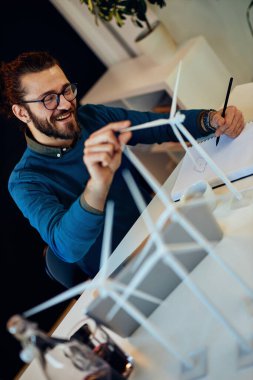 Genç yenilikçi hippi ofisinde oturuyor, yel değirmeni modeli çiziyor ve covid 19 sırasında dizüstü bilgisayarda yazıyor. Sürdürülebilir geliştirme kavramı.