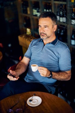 Şaraphanede oturan kıdemli yetişkin, kahve molasında kameraya bakarken akıllı telefon kullanıyor..