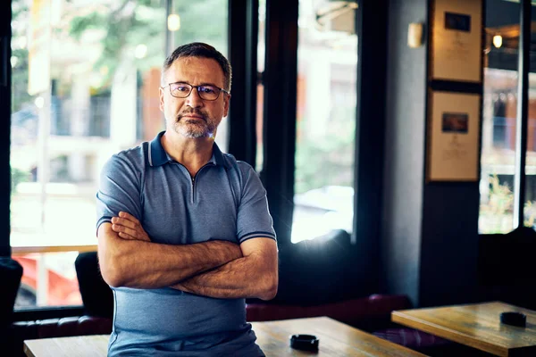Senior entrepreneur standing in his coffee shop with arms crossed and looking at camera.