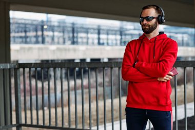 Genç, çekici, sakallı, formda bir sporcu kollarını kavuşturmuş ve şehir dışında durup müzik dinliyor..