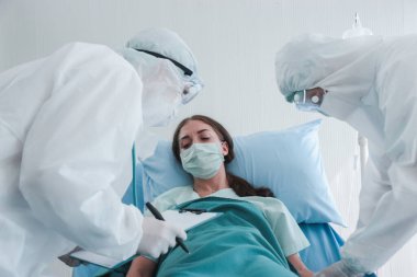 Hastalık kapmış yüz maskeli hasta kadın hastalık tedavi odasında yatakta yatıyor. Koruyucu giysiler giyen doktor, hastanedeki karantinadaki hastalarla ilgileniyor.