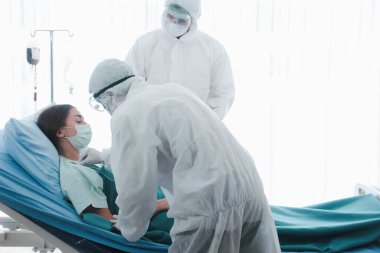 Hastalık kapmış yüz maskeli hasta kadın hastalık tedavi odasında yatakta yatıyor. Koruyucu giysiler giyen doktor, hastanedeki karantinadaki hastalarla ilgileniyor.