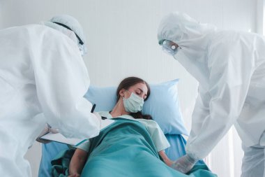 Hastalık kapmış yüz maskeli hasta kadın hastalık tedavi odasında yatakta yatıyor. Koruyucu giysiler giyen doktor, hastanedeki karantinadaki hastalarla ilgileniyor.