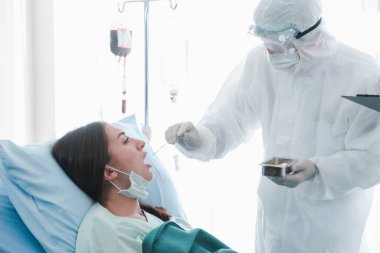 Hastalık kapmış hasta kadın, hastalık tedavi odasında yatıyor, doktor muayenesi için ağzı açık, koruyucu giysiler giyen doktor, hastanede karantinadaki hastalarla ilgileniyor.