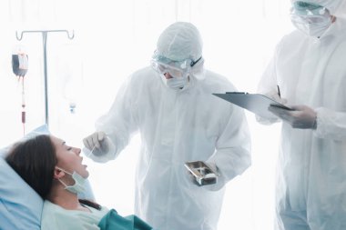 Hastalık kapmış hasta kadın, hastalık tedavi odasında yatıyor, doktor muayenesi için ağzı açık, koruyucu giysiler giyen doktor, hastanede karantinadaki hastalarla ilgileniyor.