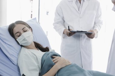 Hastalık kapmış yüz maskeli hasta kadın hastalık tedavi odasında yatakta yatıyor. Koruyucu giysiler giyen doktor, hastanedeki karantinadaki hastalarla ilgileniyor.