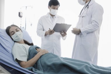 Hastalık kapmış yüz maskeli hasta kadın hastalık tedavi odasında yatakta yatıyor. Koruyucu giysiler giyen doktor, hastanedeki karantinadaki hastalarla ilgileniyor.