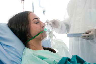 Hastalıklı hasta kadın solunum cihazlarıyla yatakta yatıyor, koruyucu giysiler giyen doktor hastalık tedavi odasındaki karantinaya bakıyor.