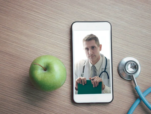 Doctor on smartphone screen, stethoscope and green apple on table, doctor online concept, online medical communication and medical consultant in nutrition on virtual interface